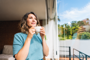 Woman with wavy shoulder-length hair holding a white coffee cup, smiling as she looks out a sunlit window toward trees and blue sky.