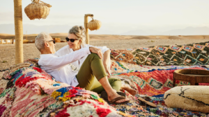 Retired couple relaxing on colourful woven rugs at a desert camp at sunset, enjoying a well-planned vacation